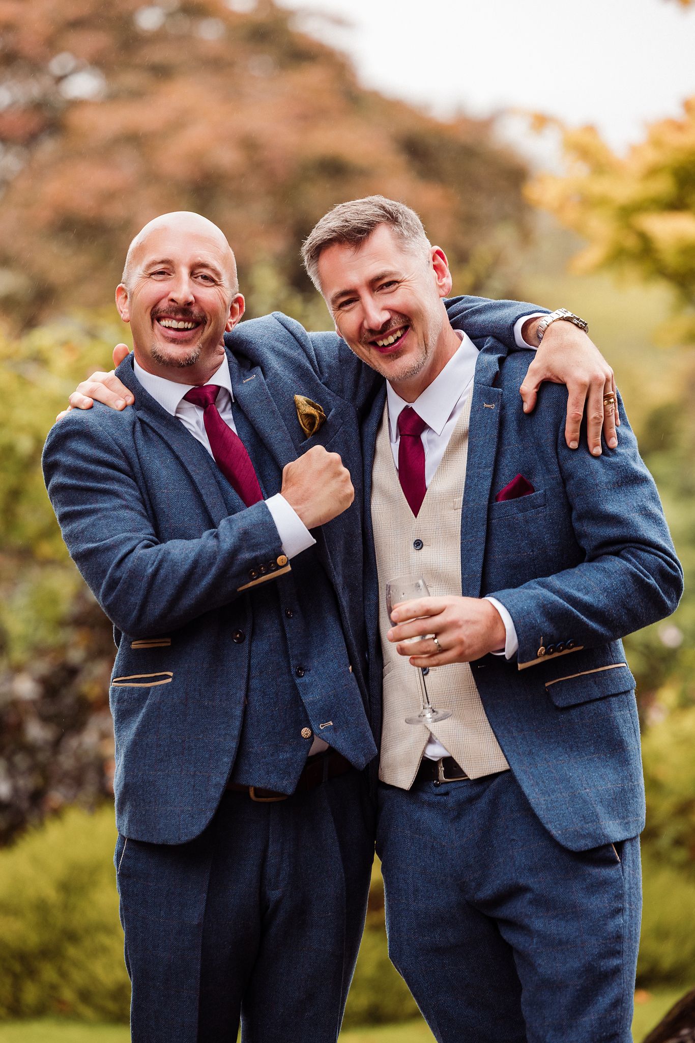 Two men in navy suits and burgundy ties share a joyful embrace and laughter during an autumn outdoor photo session.