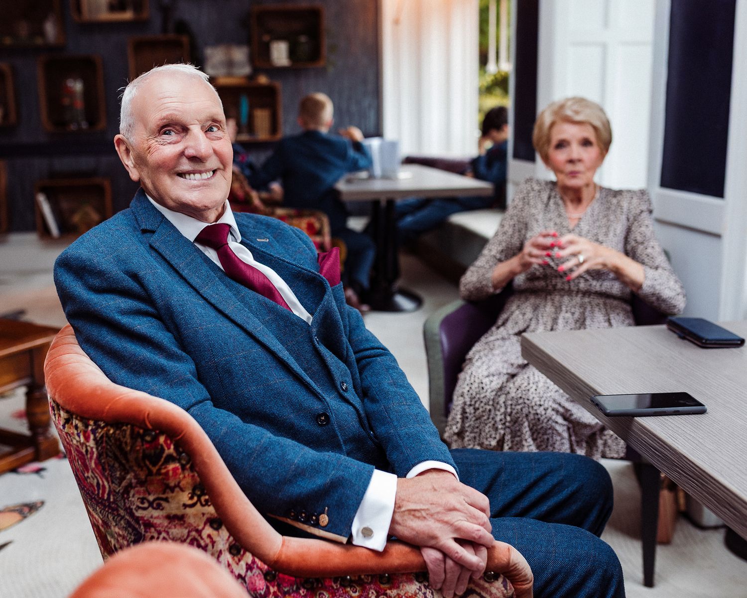 A person wearing a navy blue suit sits and smiles while conversing in an elegant indoor setting.
