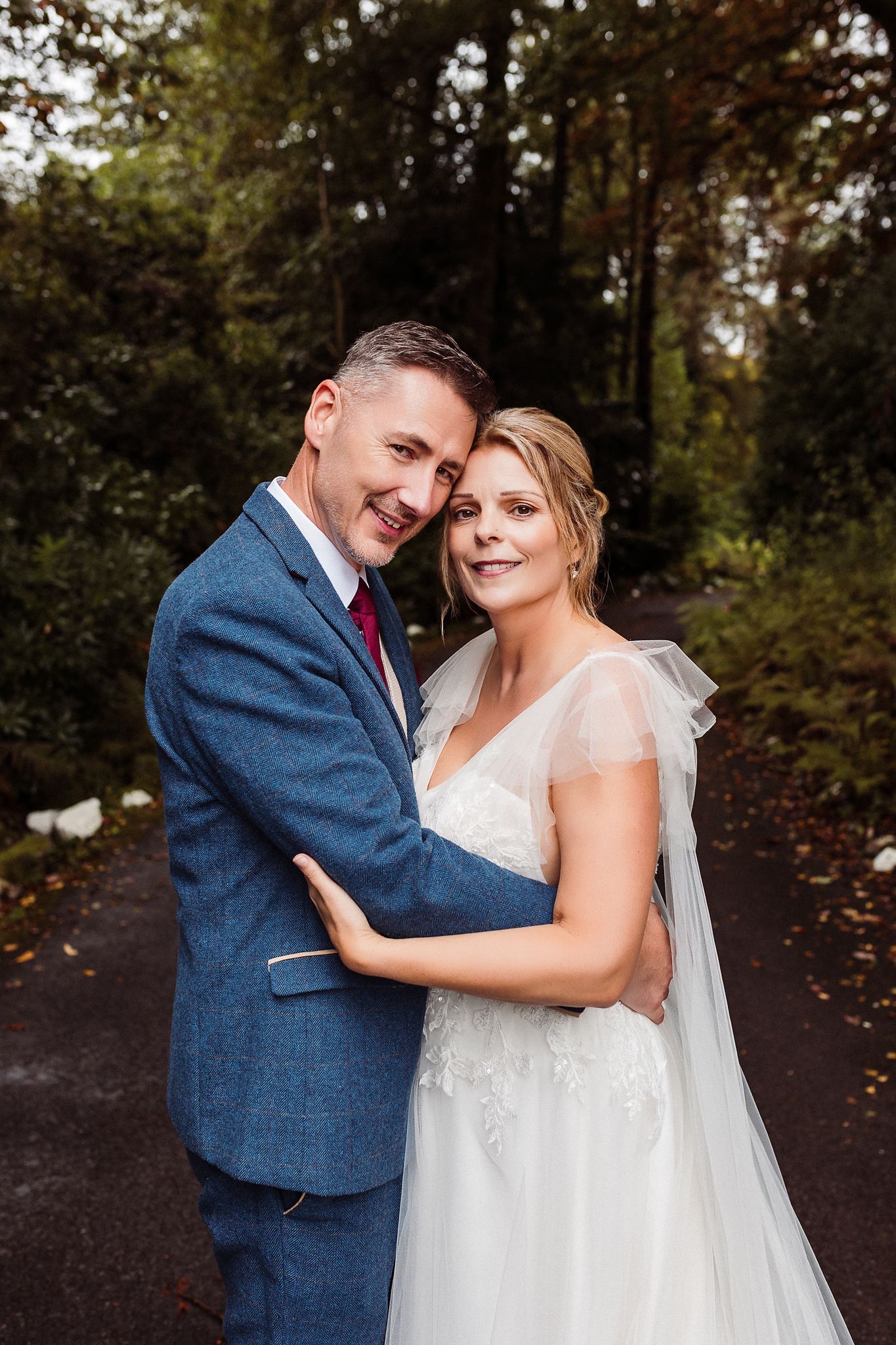 A couple in wedding attire embraces outdoors in a wooded setting with autumn foliage in the background.