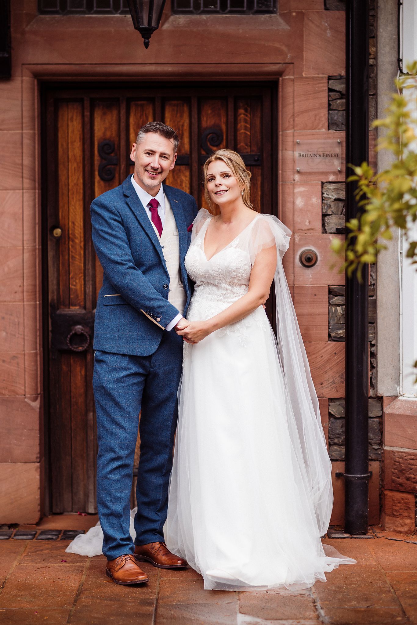 A bride and groom pose together in front of a rustic wooden door, with her in white and him in a navy blue suit.