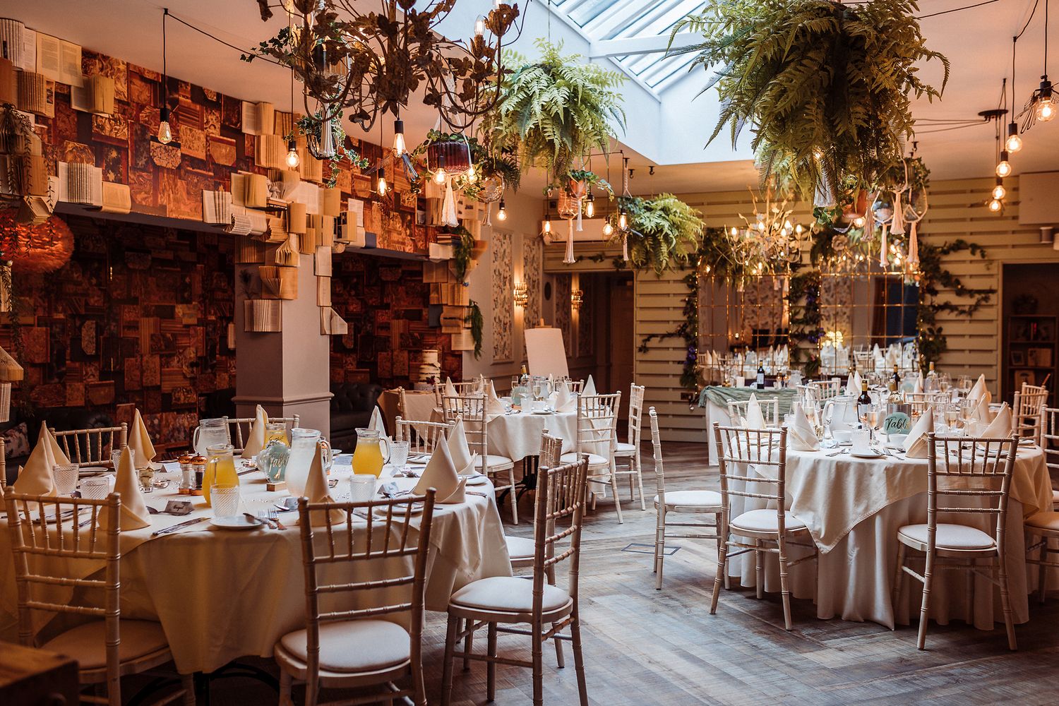 Elegant wedding reception venue with rustic decor, hanging plants, string lights and white chiavari chairs at round tables.