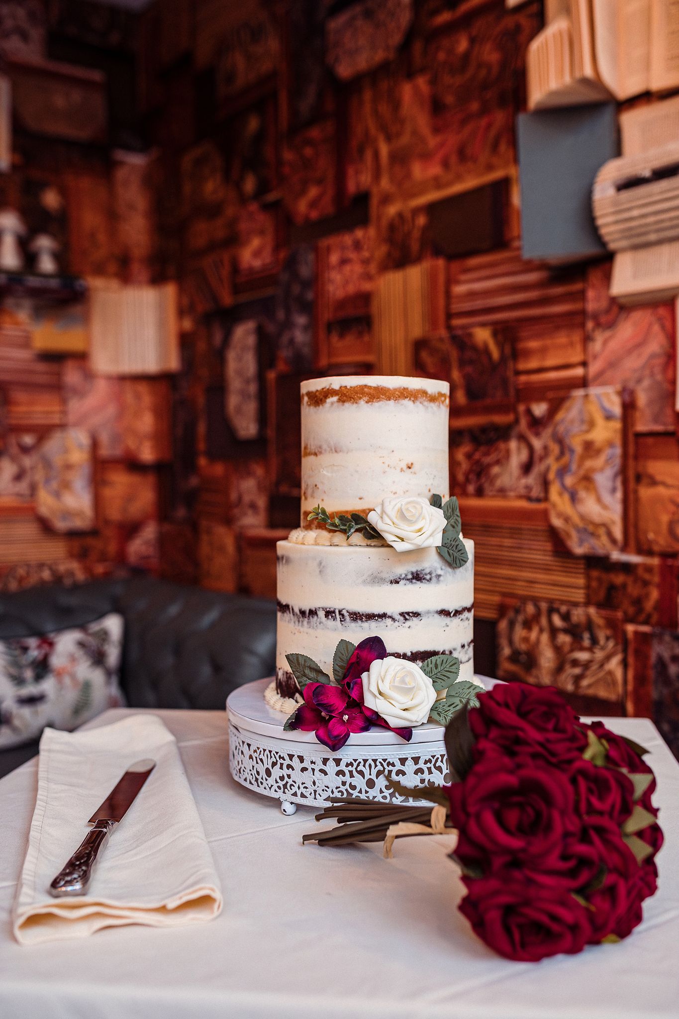 White semi-naked wedding cake decorated with deep red roses and delicate lace details on a rustic wooden table.