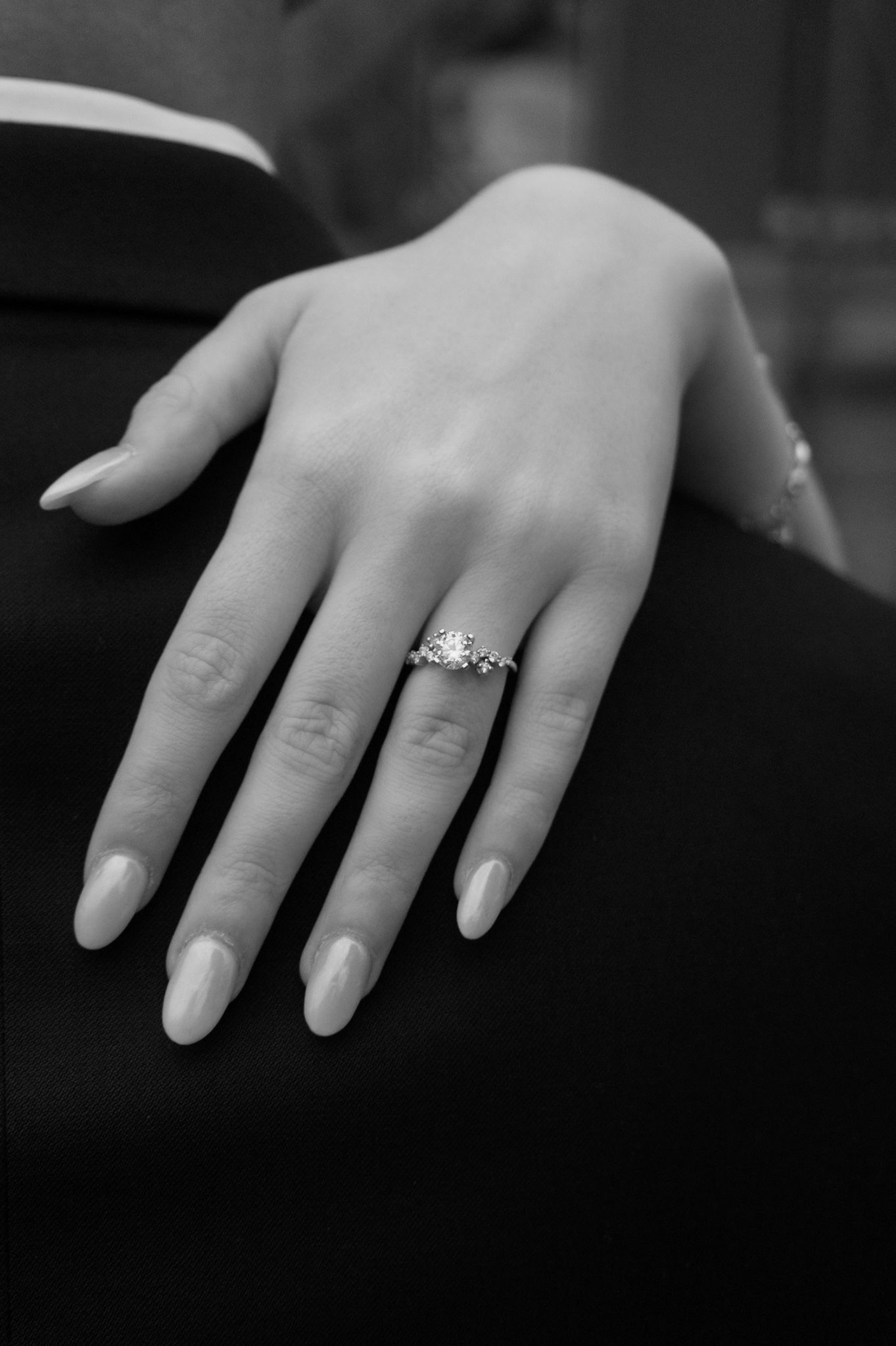 Close-up of an elegant diamond engagement ring on a woman's manicured hand against a dark background.