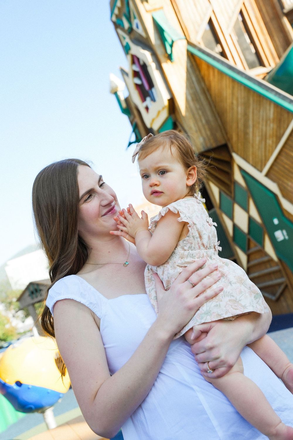 A mother in a white top shares tender moments with her baby in a floral dress outdoors.