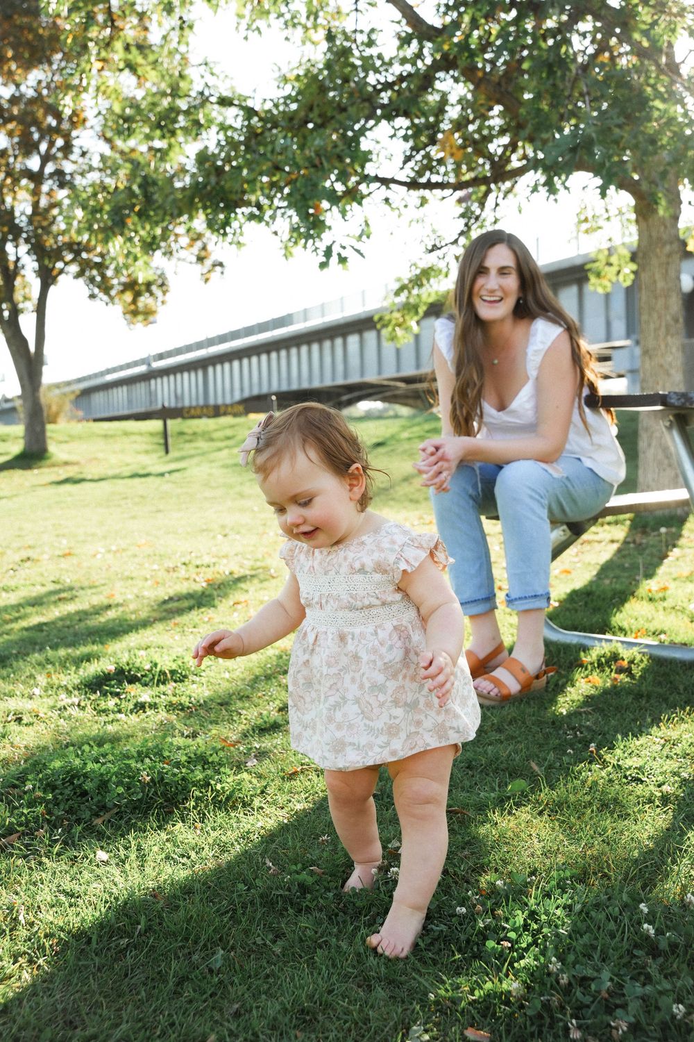 A toddler in a floral dress plays on grass near a picnic table on a sunny summer day.