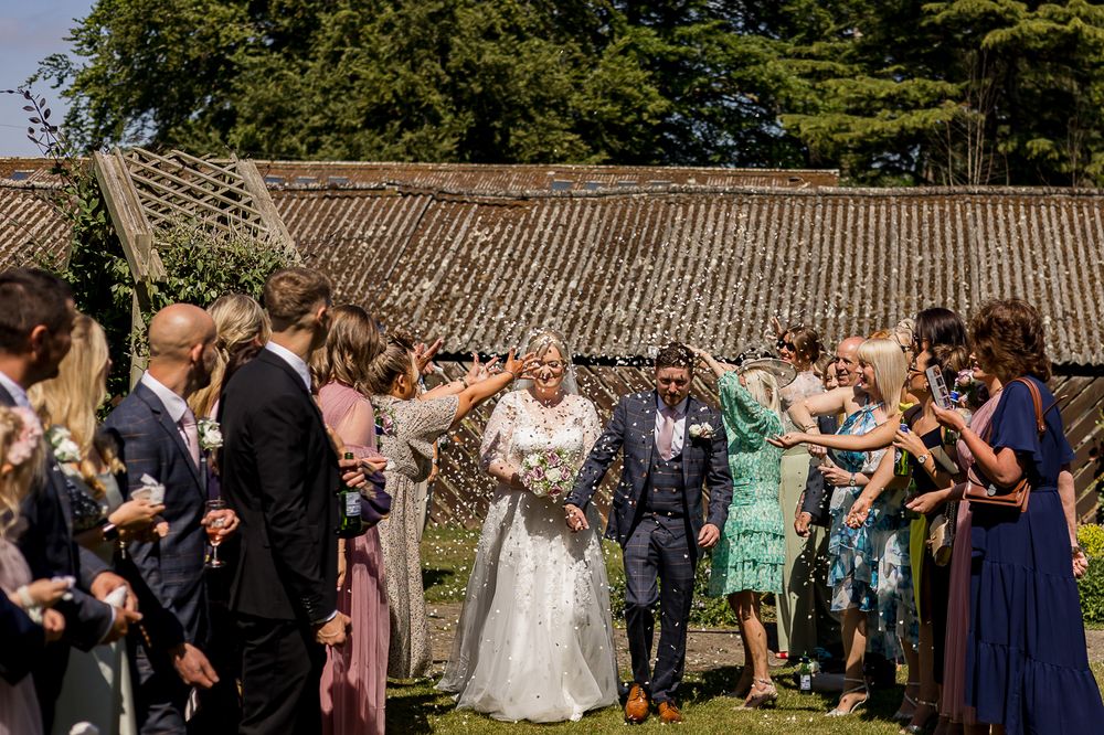 Large group of wedding guests celebrating outdoors against a rustic stone wall backdrop.