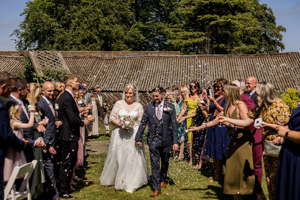 Wedding party and guests gathered for outdoor celebration near rustic stone building.