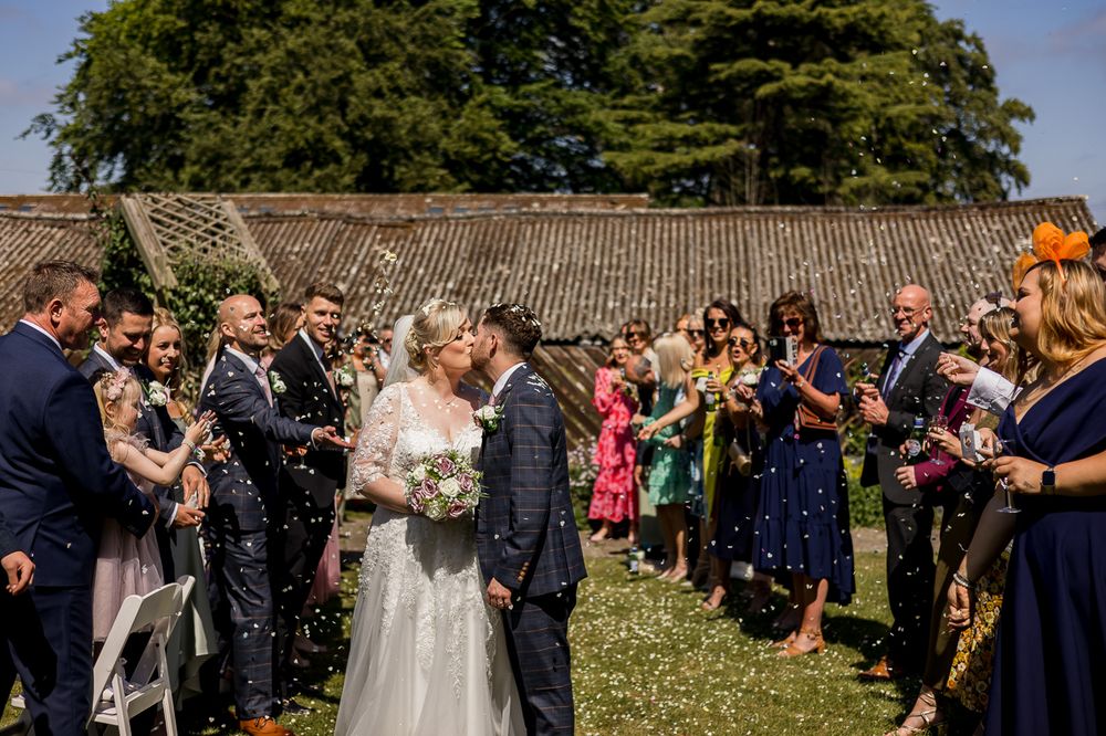 Panoramic view of wedding guests celebrating on grass near stone wall in bright sunlight.