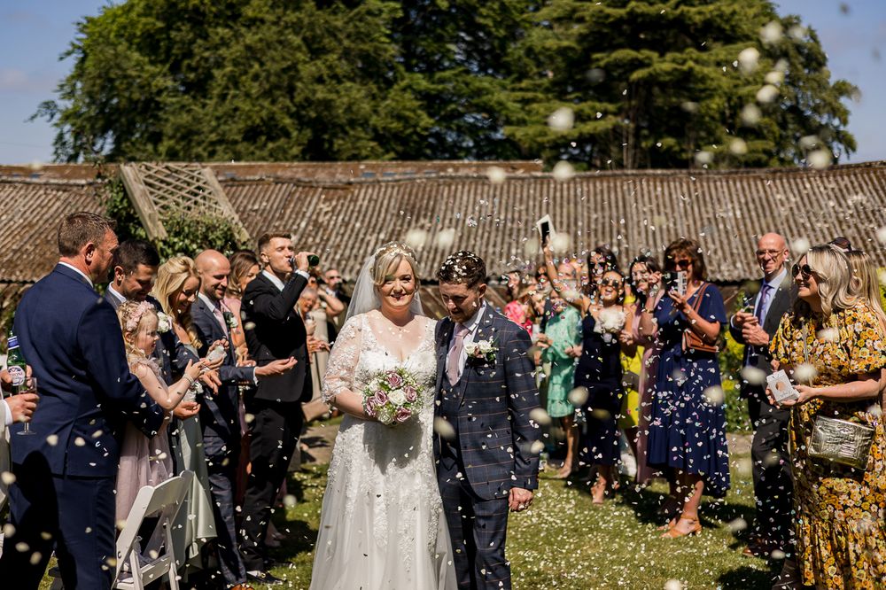 Wedding celebration panorama showing guests throwing confetti near historic stone wall.