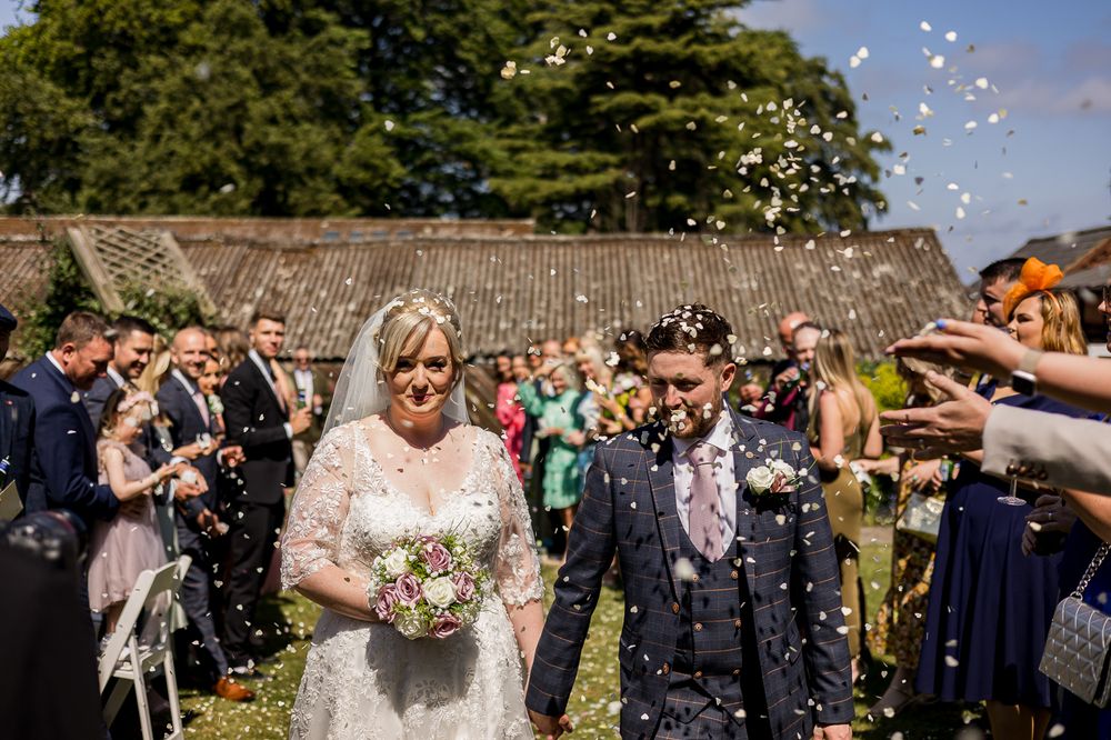 A large group of wedding guests toss confetti in a sunny garden setting.