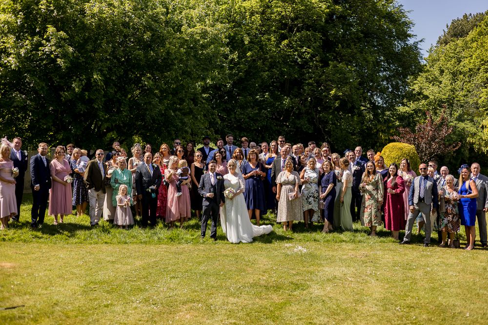 Wedding party and guests line up for a panoramic group photo on a sunny lawn.