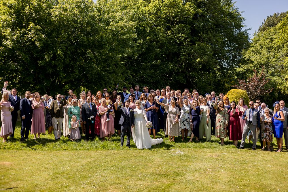 Panoramic view of wedding guests gathered on a green lawn for group photos.