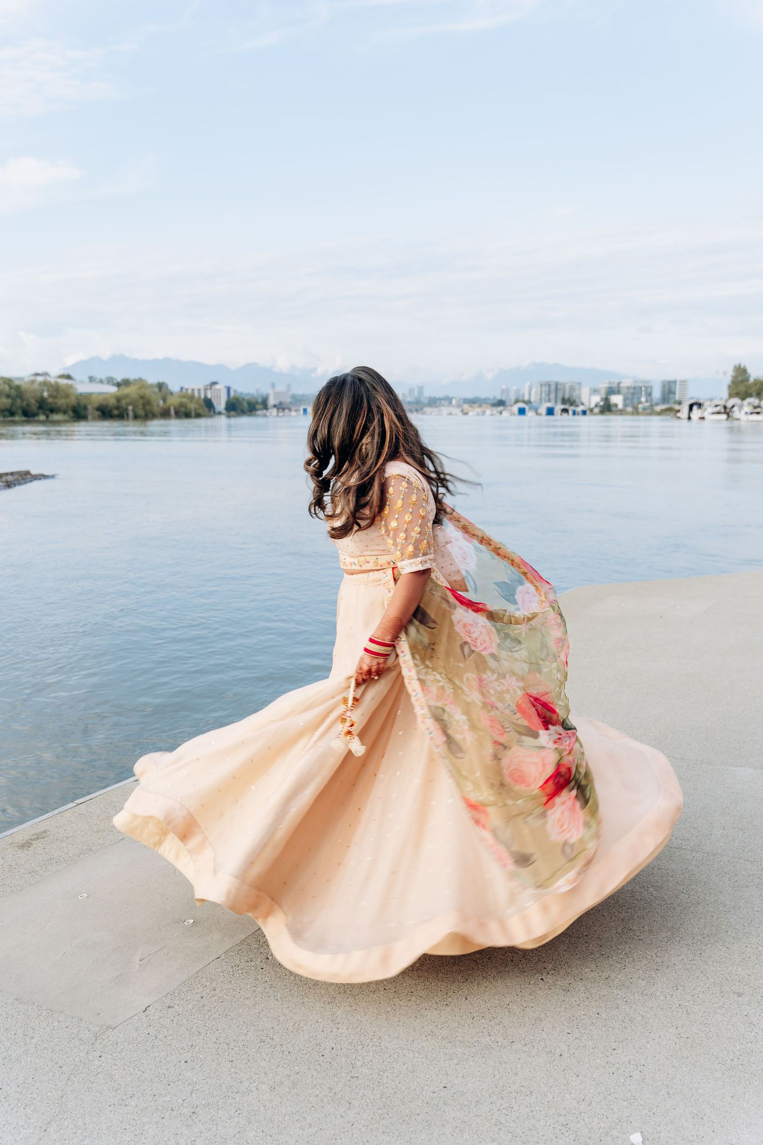 A figure in a flowing peach dress twirls gracefully on a beach with a cityscape visible in the background.