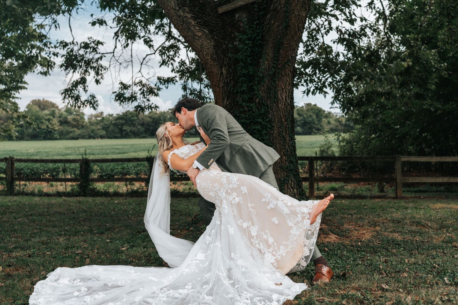 A flowing white wedding dress train billows in the breeze across green grass during an outdoor photoshoot.