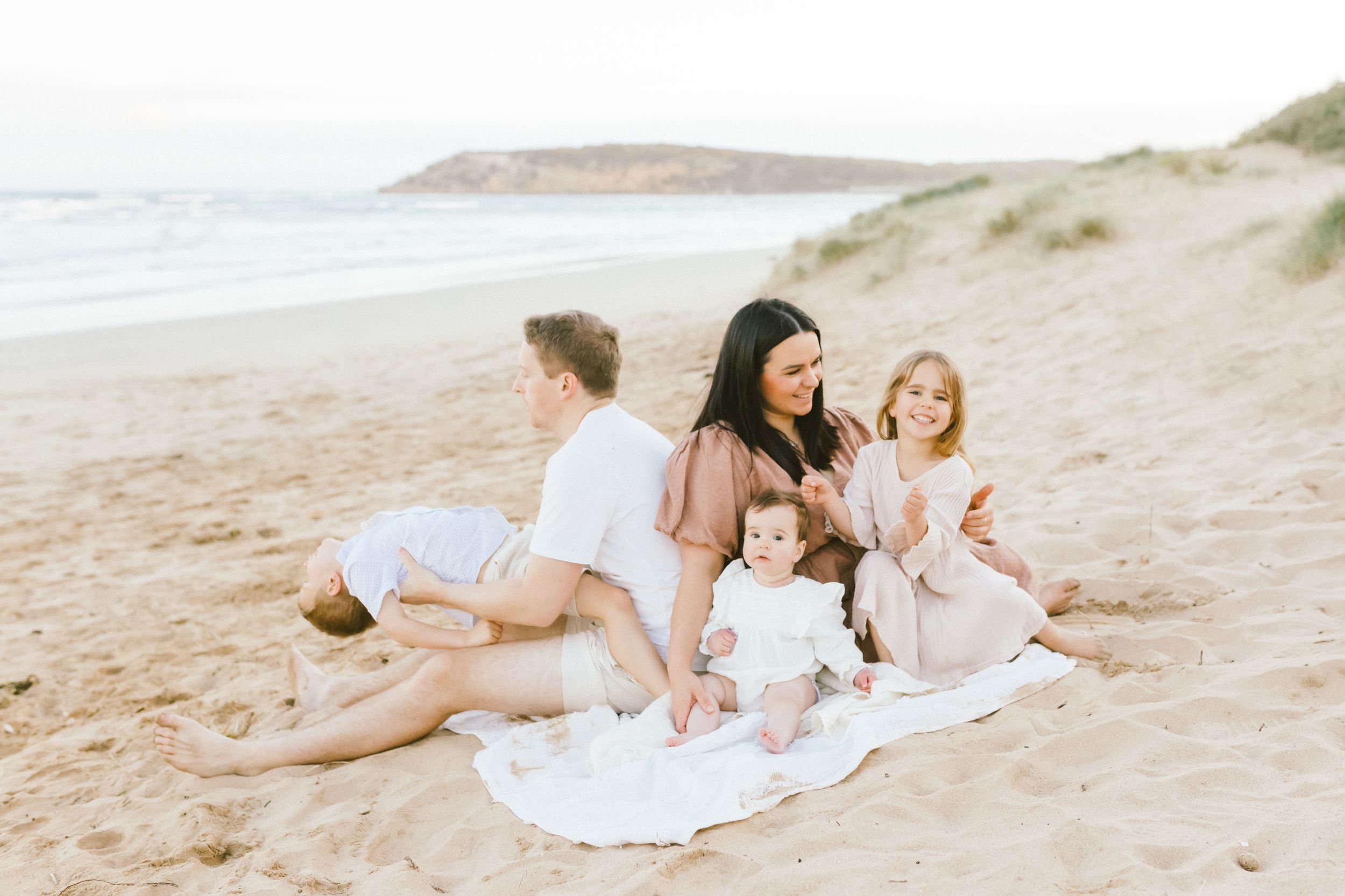A family relaxes together on a blanket at sunset on a peaceful beach with coastal cliffs in the background.