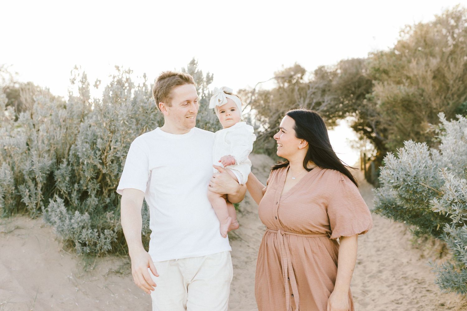 Family wearing neutral colored outfits poses together on sandy beach with coastal vegetation at sunset.
