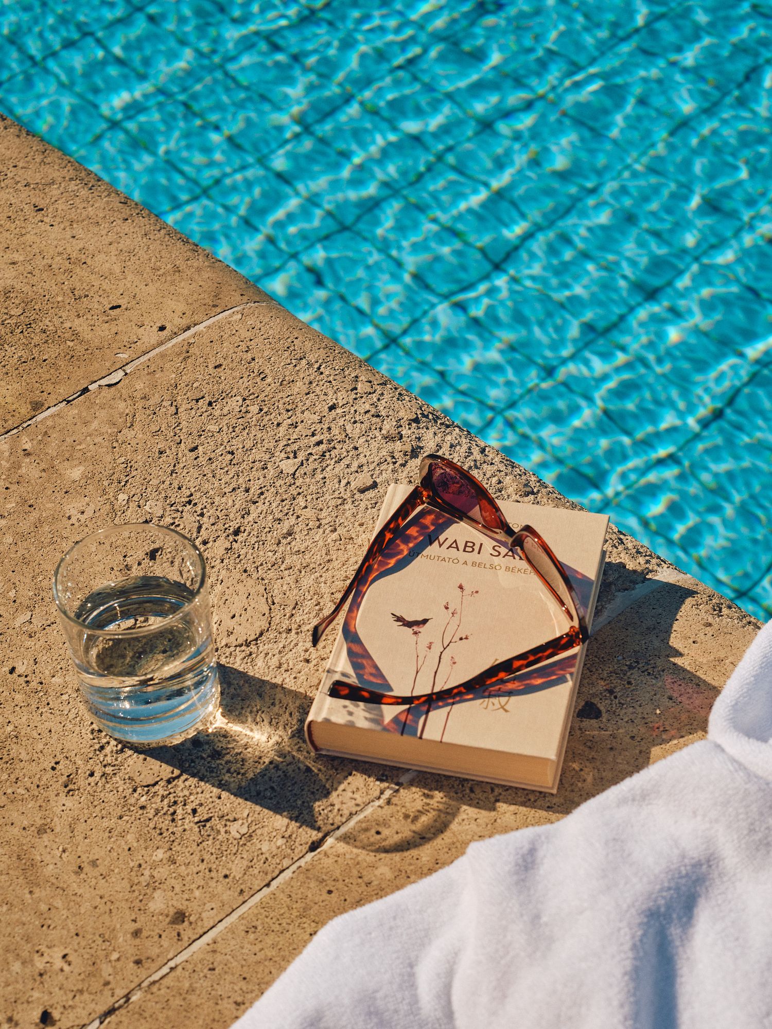 Sunglasses and wine glass casting shadows by a turquoise swimming pool on a sunny day.