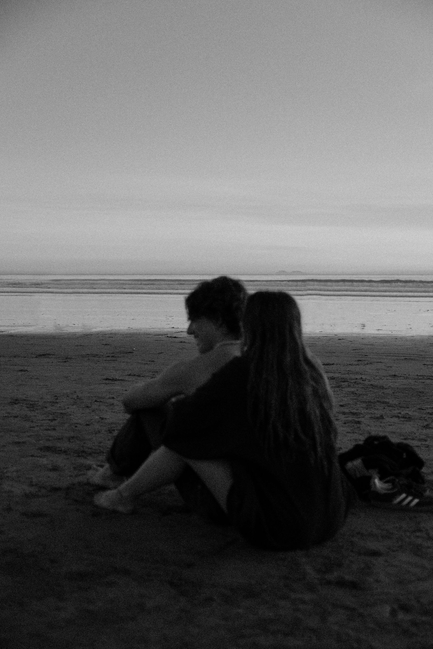 A couple sits together on a beach at sunset in black and white.