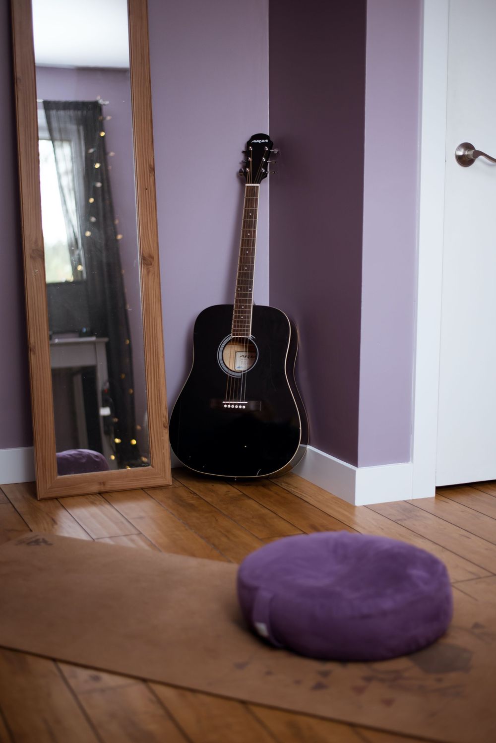 Black acoustic guitar leaning against purple wall with round purple meditation cushion on wooden floor.