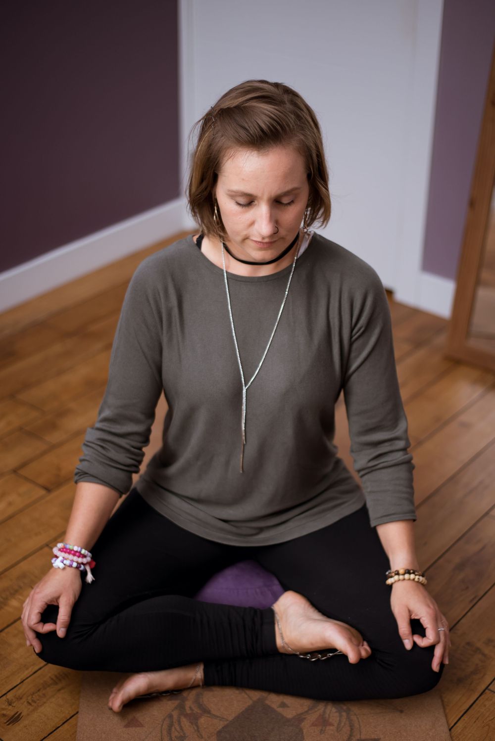 Person in meditative pose wearing brown sweater and silver necklace on hardwood floor.