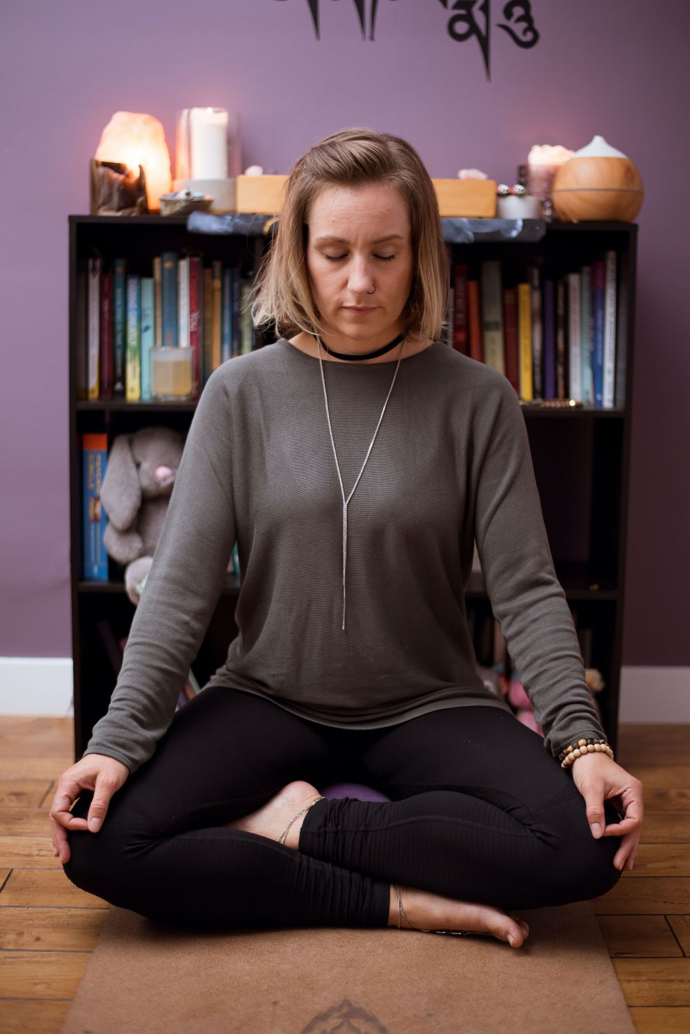 Person meditating in front of bookshelf with candles and purple wall background.