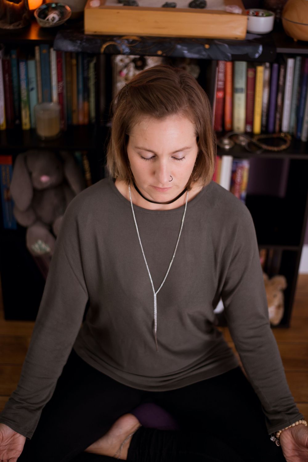 Person in meditation pose wearing brown sweater in front of bookshelf with soft lighting.