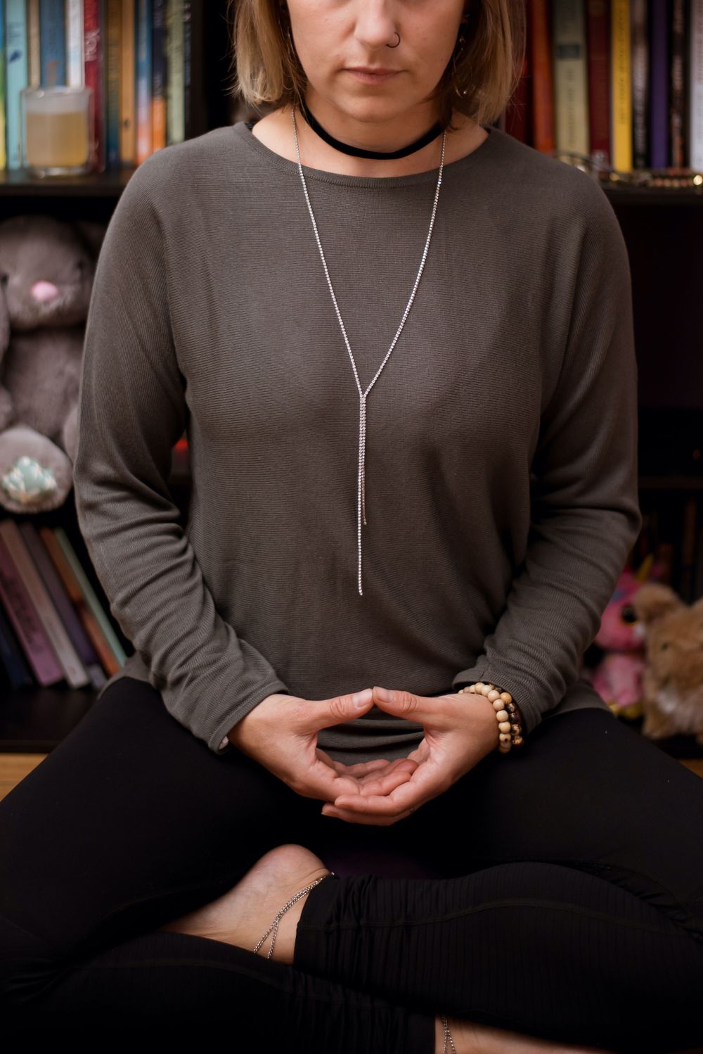 Person wearing brown sweater and silver necklace sitting cross-legged with bookshelves in background.