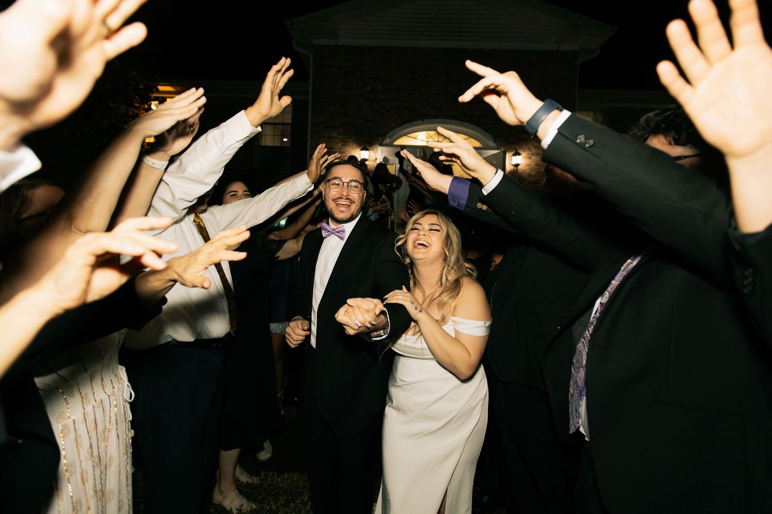 Bride and groom smile and hold hands as guests reach toward them with raised hands during a lively nighttime wedding exit.
