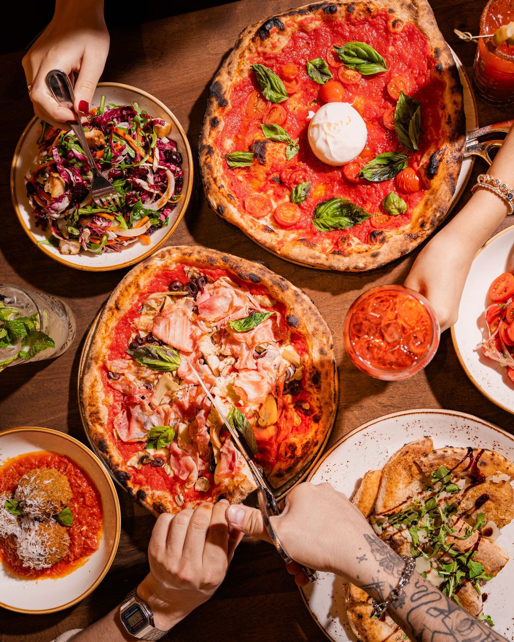 Several hands reach for Italian pizzas topped with basil and burrata alongside salads and appetizers on a wooden table.