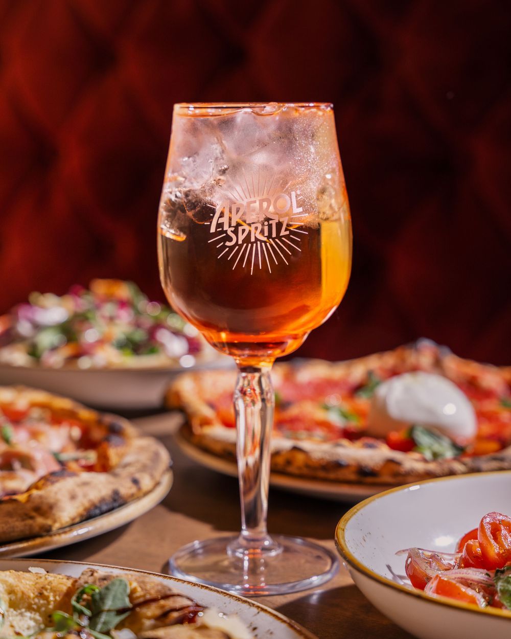 A glass of golden beer sits among plates of artisanal pizza with fresh toppings in a warm restaurant setting.
