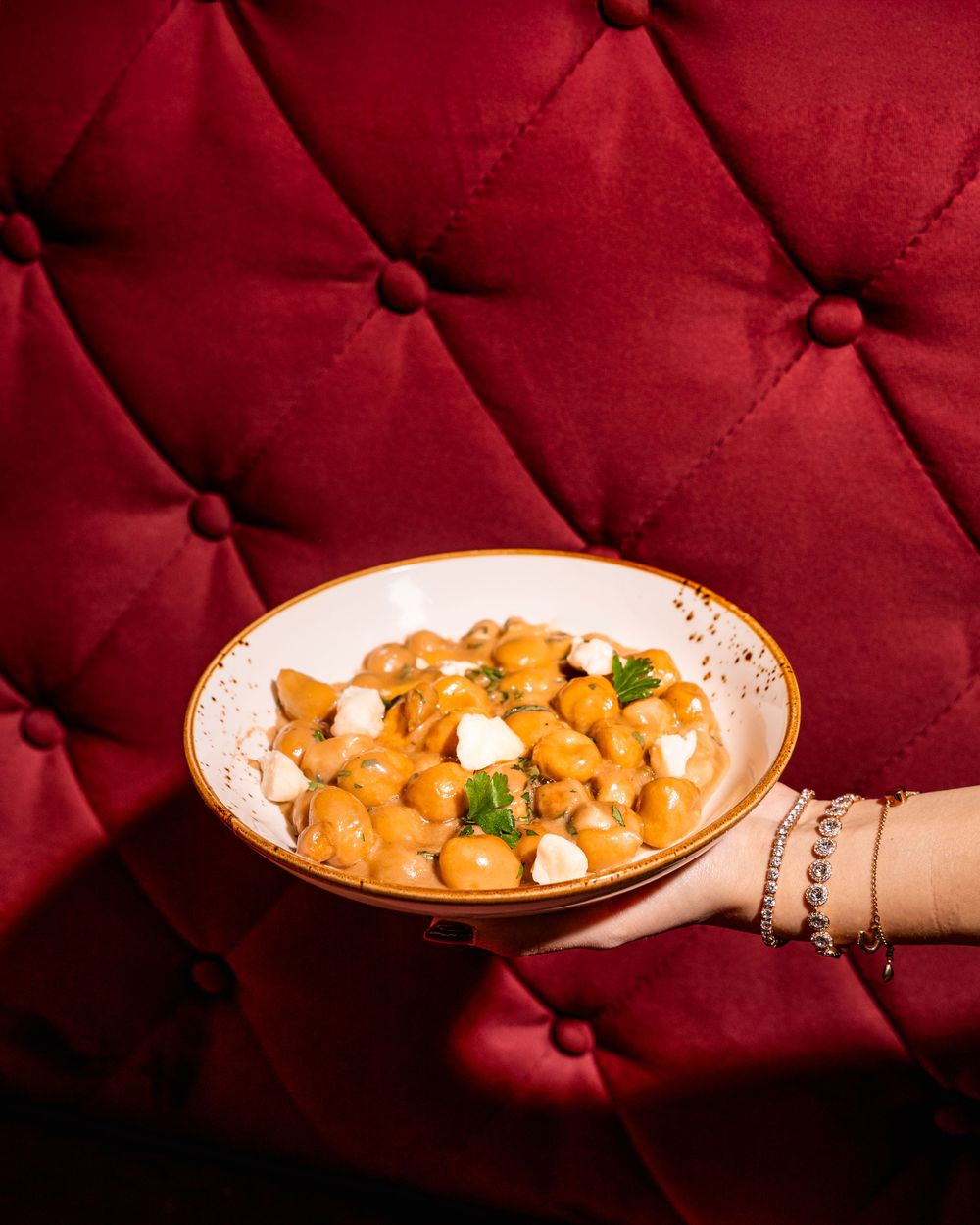 A creamy orange butter chicken curry with white yogurt dollops served in a pink bowl against a red tufted background.