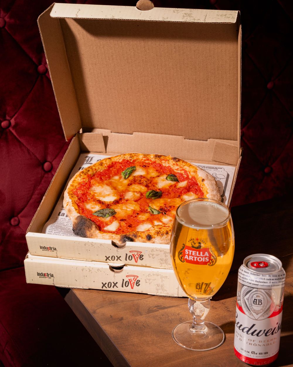 Fresh Margherita pizza in delivery box with a glass of Stella Artois beer and can on dark red background.