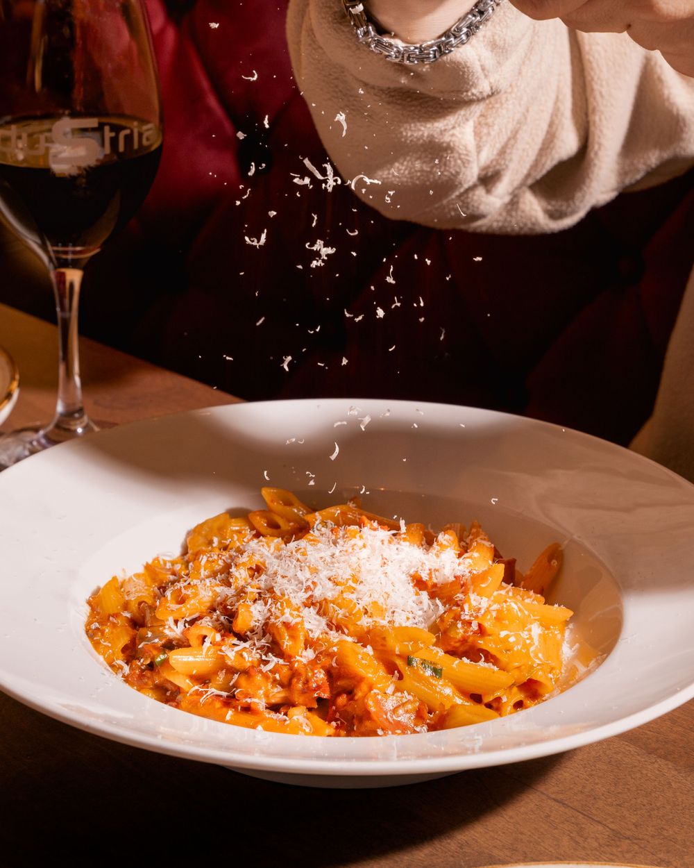 Parmesan cheese being sprinkled over a plate of orange pasta in tomato sauce with a wine glass in the background.
