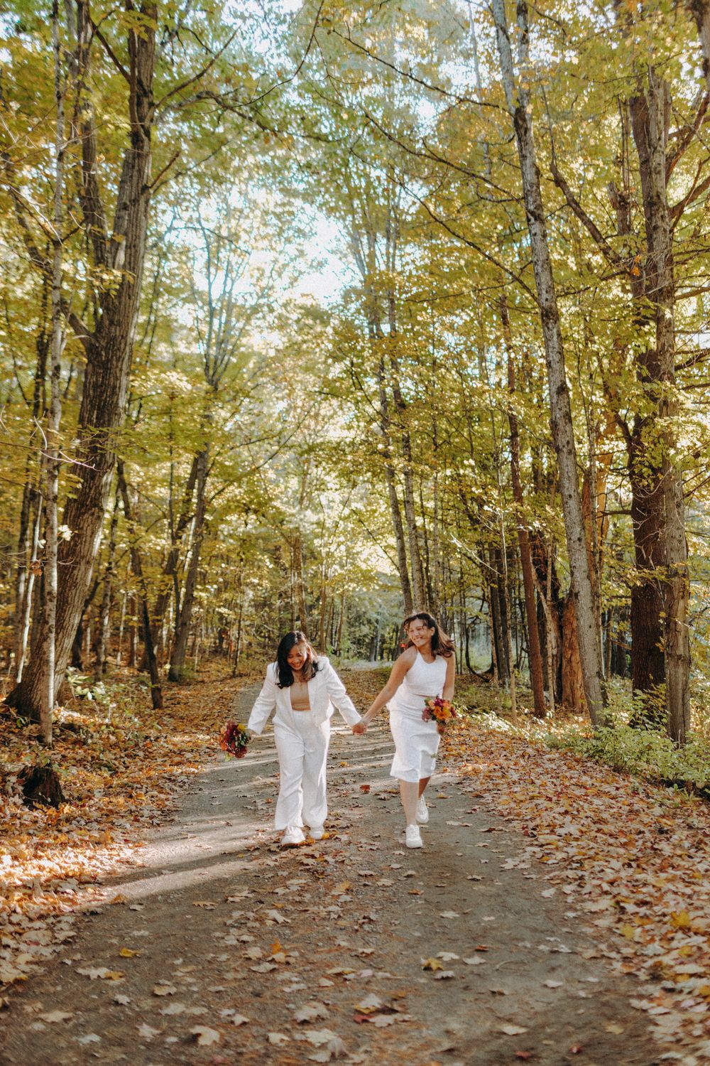 Wedding party walking through a tree-lined path covered in fallen autumn leaves.