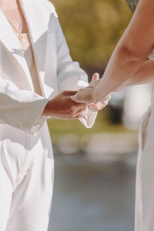 Close-up details of white wedding dresses and hands during an outdoor ceremony by the water.