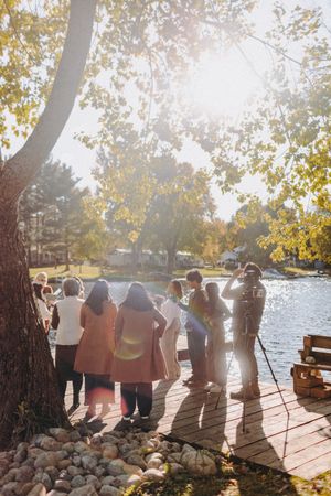 Wedding guests gathered on a wooden dock during golden hour with sunlight filtering through autumn trees.