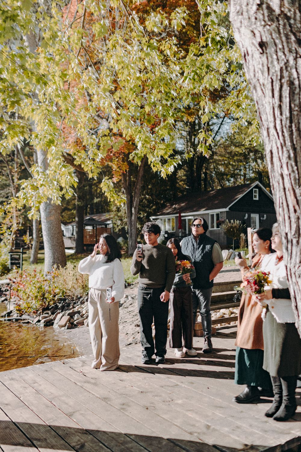 Wedding party standing on a wooden boardwalk surrounded by fall foliage near a lake.
