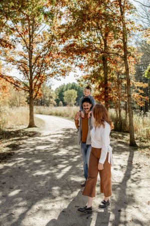 Lifestyle family photographer capturing parents cuddling toddler son on shoulders in sunlit autumn forest