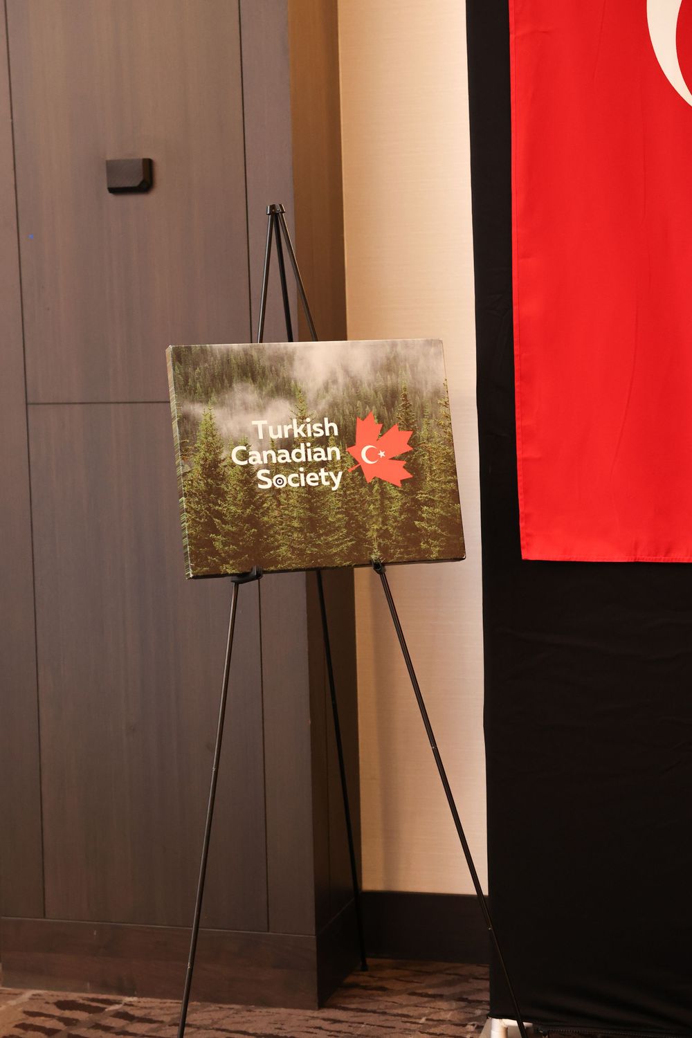 Turkish Canadian Society sign with maple leaf logo displayed on an easel next to red flag.