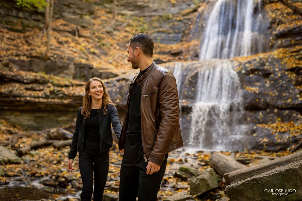 A couple poses together near a multi-tiered waterfall surrounded by golden autumn leaves.