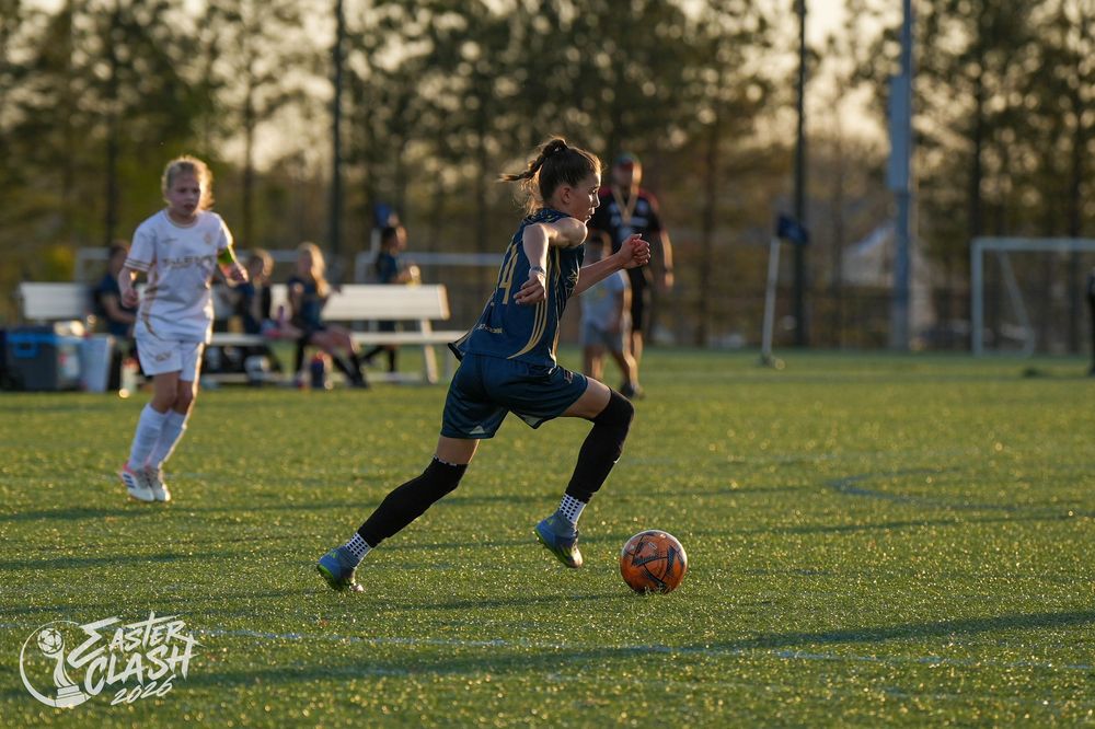Female soccer player dribbles ball on green field during Easter Clash tournament game at sunset.