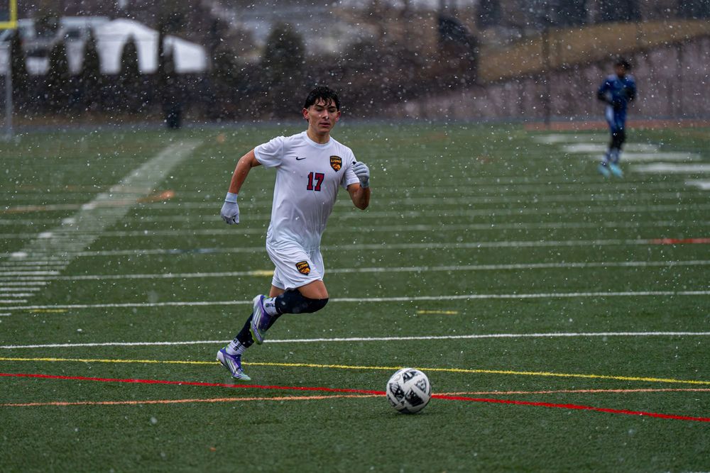 Soccer player in white jersey runs across field during match while opponent follows in blue uniform.