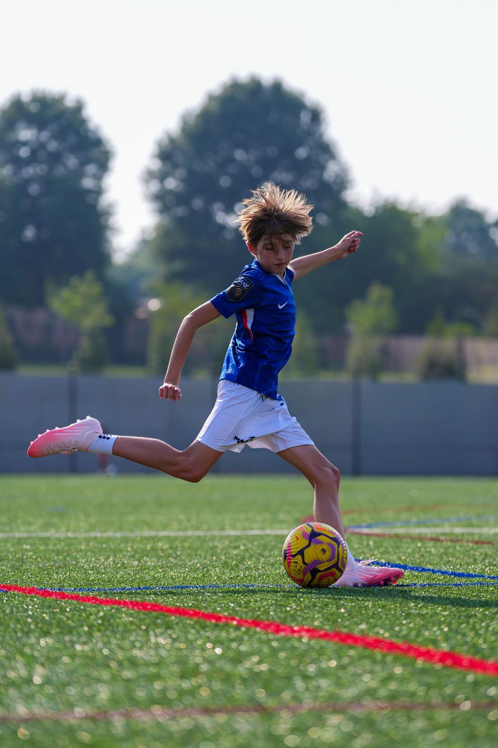 Young soccer player in blue jersey kicking a yellow ball on a green turf field during a sunny day.