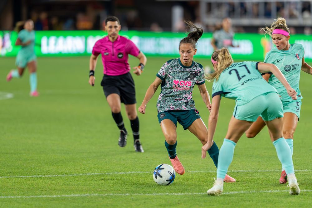 Female soccer players compete for the ball during a night match, with a referee watching closely in the background.