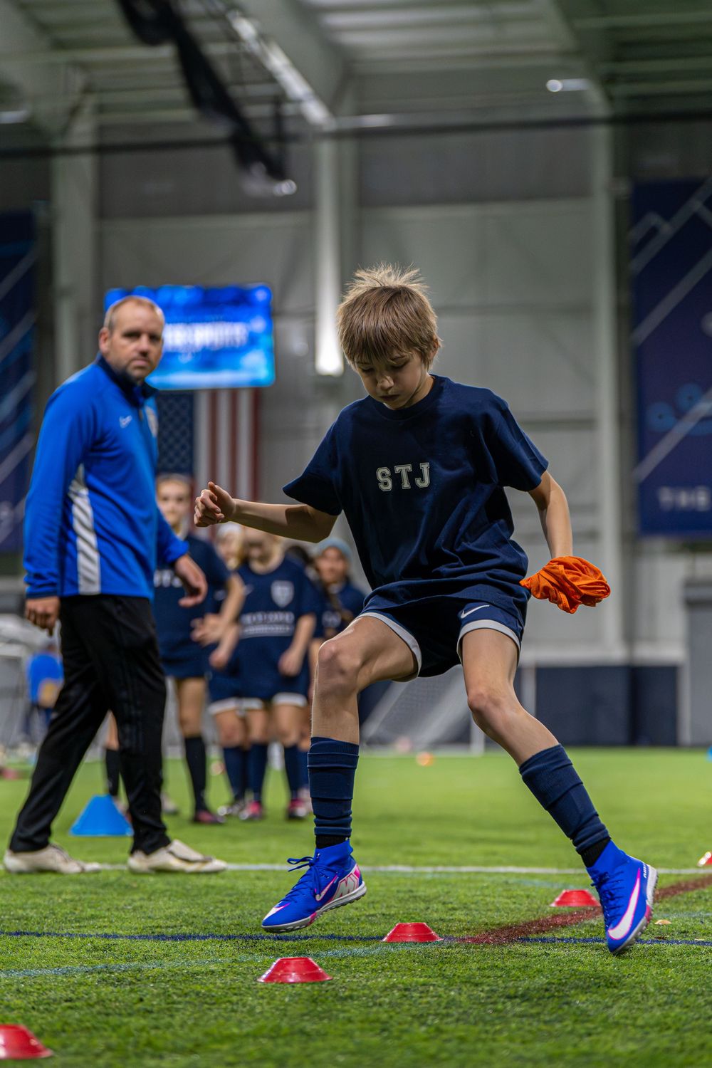 Soccer players practice footwork drills between red cones on an indoor turf field during training session.