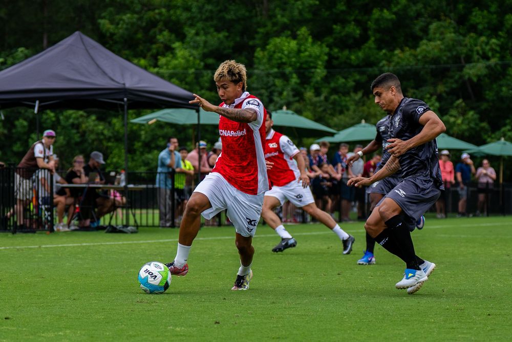 Soccer player in red and white Arsenal kit dribbles ball past defender in outdoor match.