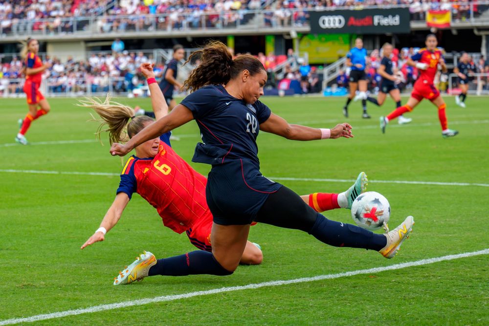 Two women's soccer players slide tackle during an intense match at Audi Field stadium.