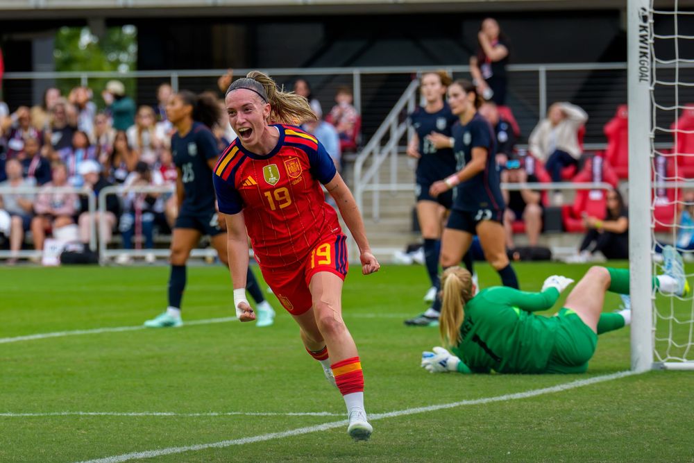 Spanish female soccer player #19 celebrates a goal in red uniform as goalkeeper lies on ground during a match.