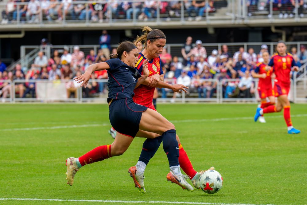 Two female soccer players compete for the ball during a match on a green field with fans in the stadium.