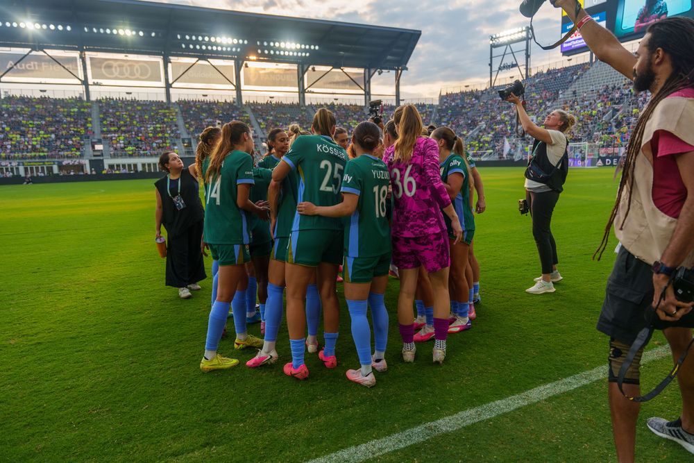 Women's soccer team huddles on field in green uniforms before match in packed stadium.